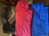 Three jackets laid flat: Pink The North Face hooded jacket XXL, Blue The North Face collar jacket small, Black Chic ET Jeune jacket with leatherette body and multicolored sleeves.