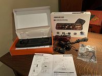 Photo showing the streaming media rearview mirror inside its packaging box along with cables, mounts, connectors and user manual spread out on a table.