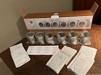 Photo of six aluminum magnetic canisters placed in a wooden engraved rack, unopened spice pouches, and printed labels displayed on a wooden surface.