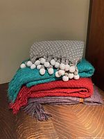 Stack of four throws: grey/white cotton throw with pom-poms on top, teal, red, and purple acrylic throws below, all folded.