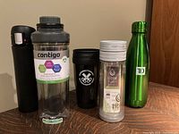 Front view of five travel cups and bottles on wooden surface: black insulated tumbler, Contigo shaker cup with label, black tumbler with white logo, Libre glass travel mug, green stainless steel water bottle
