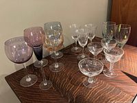 Photo showing the full assortment of wine glasses described by the seller on a wooden surface against a neutral background. Includes multi-coloured, etched, recycled, and plain glass varieties.
