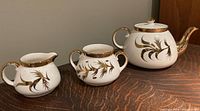 Front angle view of vintage tea set showing teapot, creamer, and sugar bowl with gold leaf decoration on white porcelain surface placed on wooden surface.