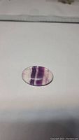 Natural oval cabochon fluorite gemstone shown on a flat light background, highlighting its purple banding and smooth polished surface.