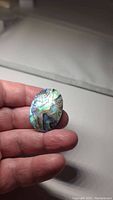 Natural AAA abalone doublet gemstone held between fingers, showing oval cabochon shape with vivid purple, blue, green iridescence.
