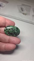 Oval green zoisite gemstone with red ruby inclusions, held between fingers for scale, showing polished smooth surface and natural color patterns.