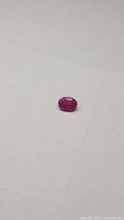 Close-up photo of a small oval natural ruby placed on a plain white background, showcasing the stone's color and polish.