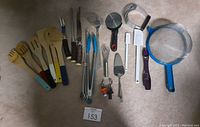 Photo showing all kitchen utensils in a group on beige carpet, including wooden utensils with colored handles, meat carving set, skewers, tongs, masher, cork replacements, mini whisk, hand strainer, pizza roller, scrapers, dough kneader and spice spoon.