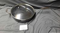 Epicure frying pan with glass lid set on a gray fabric background, visible frying pan body and lid handle.