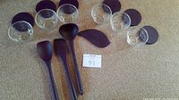 Six small clear glass prep bowls with black silicone lids arranged in a row, with four black kitchen utensils including a ladle, spoon, sauté spoon, and pan scraper laid in front on a beige carpet.
