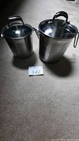 Two stainless steel strainer pots, one smaller and one larger, with glass lids on carpeted floor.