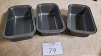 Photo showing three dark gray metal loaf pans arranged side by side on carpet. Each pan is rectangular with slightly flared edges and smooth interiors, consistent with typical non-stick bakeware.