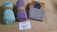 Two rolled Norwex kitchen towels in mint and purple, accompanied by a branded Norwex box containing two Spiri sponges and two metallic-textured sponges beside the box, all placed on a beige surface.