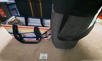The multicolor striped Large Utility Tote with black handles and insulated interior next to the grey and black Stand Tall Bin on carpeted floor.