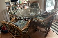 Full view of round glass top rattan kitchen table with four swivel chairs arranged around it. Cushions are on all chairs.