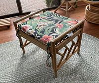 Photo shows a light-colored rattan ottoman with a removable cushion tied at the corners. The cushion has a tropical floral print.