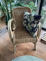 Small rattan chair placed on a hardwood floor near a plant, showing woven seat, backrest and armrests of rattan material.