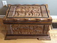 Front view of African wooden chest showing carved rhinoceros design on front panel with smaller animals and patterned side borders. Chest has scalloped edges and raised base.