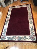 Full view of the rectangular area rug laid on wood flooring, showing the burgundy center and detailed floral border with fringes on one end.
