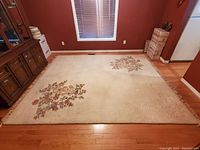Top-down view of the full wool area rug placed on wooden flooring showing two floral clustered designs on a light beige background and evidence of wear and dirt on one section.