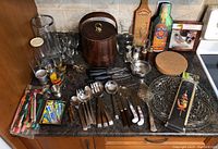 Photo showing assorted bar entertaining items on countertop: large ice bucket, glassware including champagne flutes and shot glasses, wooden container, bottle openers, stir sticks, and serving utensils.