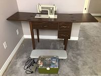 Kenmore sewing machine built into wooden cabinet with fold-out wings; foot pedal and green accessory box on the floor