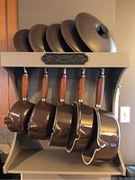 Front view of five brown sauce pots with lids hanging from wooden stand