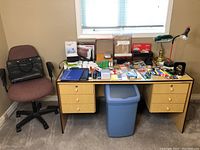 Wide view of the office lot showing a burgundy swivel chair, office desk with various office supplies on top, a blue plastic wastebasket under the desk, and two lamps on the desk.