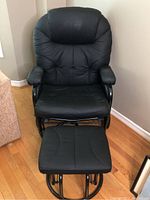 Front view of black leather rocker chair with padded armrests and backrest, along with matching ottoman in front.