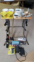 Full overview of lot showing Black & Decker workmate, plastic containers with nails, stud locator tool, hot air paint remover tool, converters, and other items on carpeted floor.