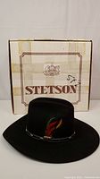 Photo of black Stetson hat with colorful feather on band in front of original Stetson box showing brand and condition.