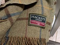 Close-up image of Hackett London anniversary tweed wool throw blanket with label dated 1988 showing plaid pattern in tan with blue and brown stripes and fringes.
