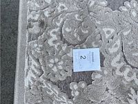Photo showing the beige rug with vintage lace floral pattern and textured plush fabric, with a label indicating lot 2