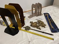Overview of all items showing six metal book ends in different colors, two brass door knockers, brass window fastener, and brass fire poker next to measuring tape