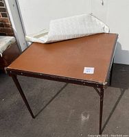 Square folding card table with brown top and metal legs, showing wear and scuffs. Padded white quilted cover partially pulled back revealing table surface.