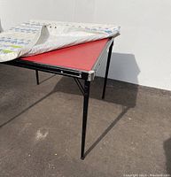 Image showing the card table partially covered with the padded cover, revealing the red top and metal legs.