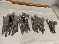 Various stainless steel cutlery pieces including knives, forks, and spoons arranged in separate piles on a white cloth surface.