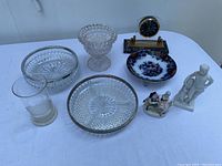 Photo shows full view of the lot on a white surface including glassware, porcelain figurines, china plate, and clock.