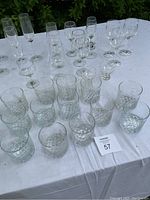 Top-down image showing glassware assortment on white cloth table cover, including wine glasses, champagne flutes, and smaller glasses arranged in groups.