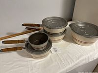 Photo showing five cream-colored cast aluminum pots nested together, revealing interior and wooden handles, on a white cloth background.