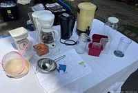 Photo of various kitchen small appliances and utensils on a white tablecloth outside. Includes coffee makers, kitchen scale, jars, containers, strainer and other items.