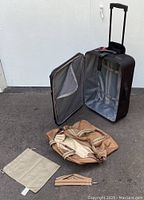 Black expandable suitcase open and standing upright with tan carry bag, beige shoe bag, and small matching pouch in foreground on concrete ground.