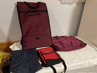 Photo showing four bags: a burgundy carry bag with collapsible wheels, burgundy hold-all bag, red and black lunch bag, and blue shoe bag.