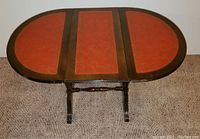 Solid wood drop leaf table fully extended showing oval red inlay surface with wood border and carved legs.