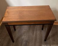 Top view of the wooden piano bench showing the flat surface and four straight legs on a wood floor.