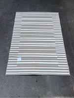 Top-down photo showing entire rectangular beige rug with white horizontal stripes laid flat on pavement.