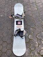 Photo showing top side of white snowboard with attached broken bindings, lying on pavement.