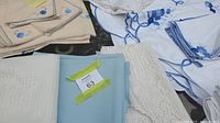 Overview of assorted table linens including cream and white embossed fabrics, crocheted lace, and light blue cloths with a tag labeled '63'.