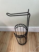 Photo of a freestanding bronze-colored metal toilet paper holder with an arm to hold one roll and a basket below for storing extra rolls. Positioned against a white wall on a hardwood floor.