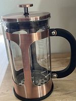 Photo showing French Press coffeemaker with copper metal casing, glass body with measurement markings, and black handle on wood surface.